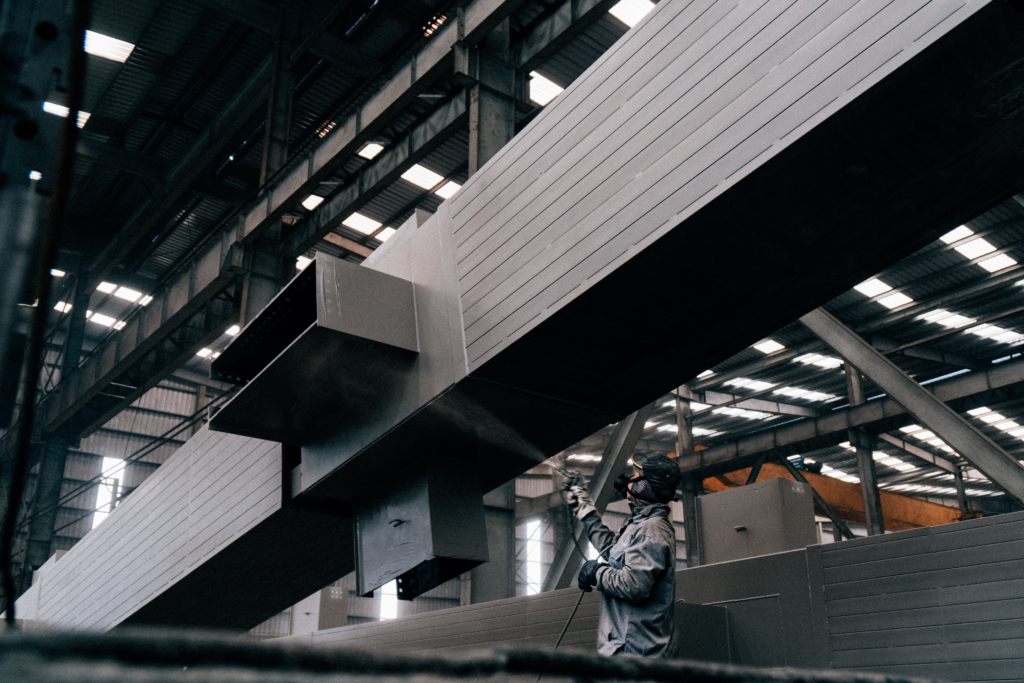 A worker sprays paint on large metal beams in an industrial warehouse.