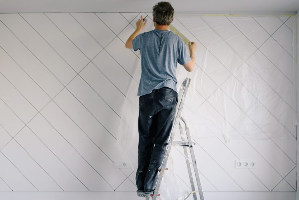 Full body back view of faceless male in shirt and dirty pants repainting wall while standing on ladder in bright empty room in daytime