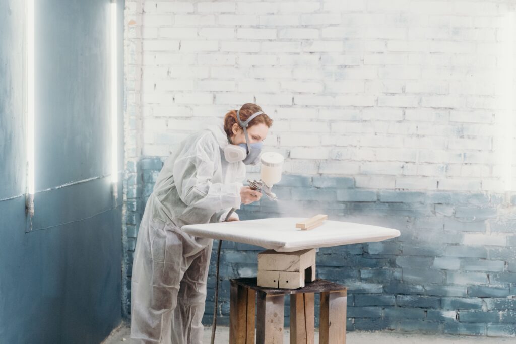 Woman wearing protective gear spray painting a canvas in an indoor art studio.